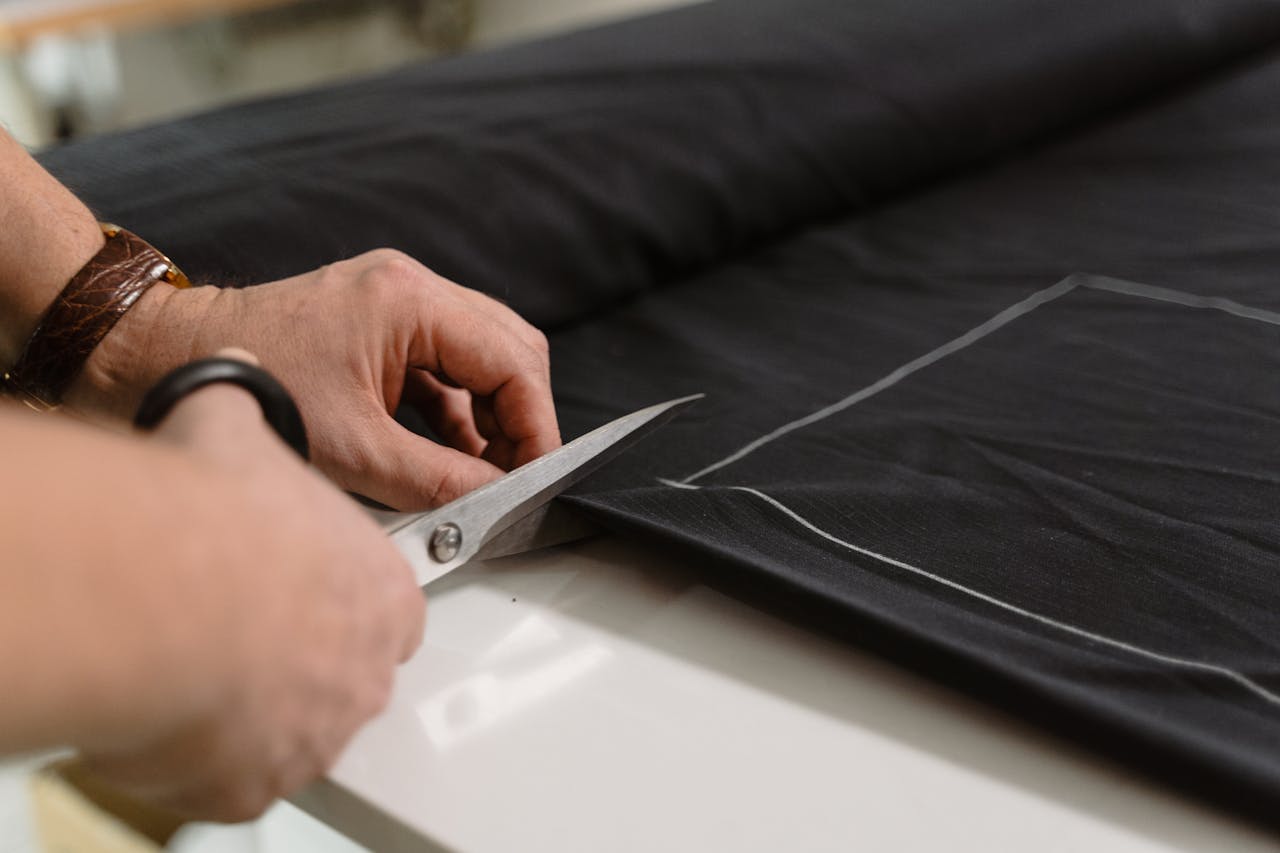 heros-img Close-up of a tailor's hand cutting black fabric with scissors, highlighting craftsmanship.